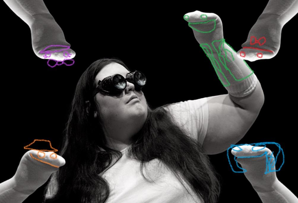 A  female-presenting gender fluid person, wearing aviator glasses, in the midst of sock puppets with lines drawn on their faces.