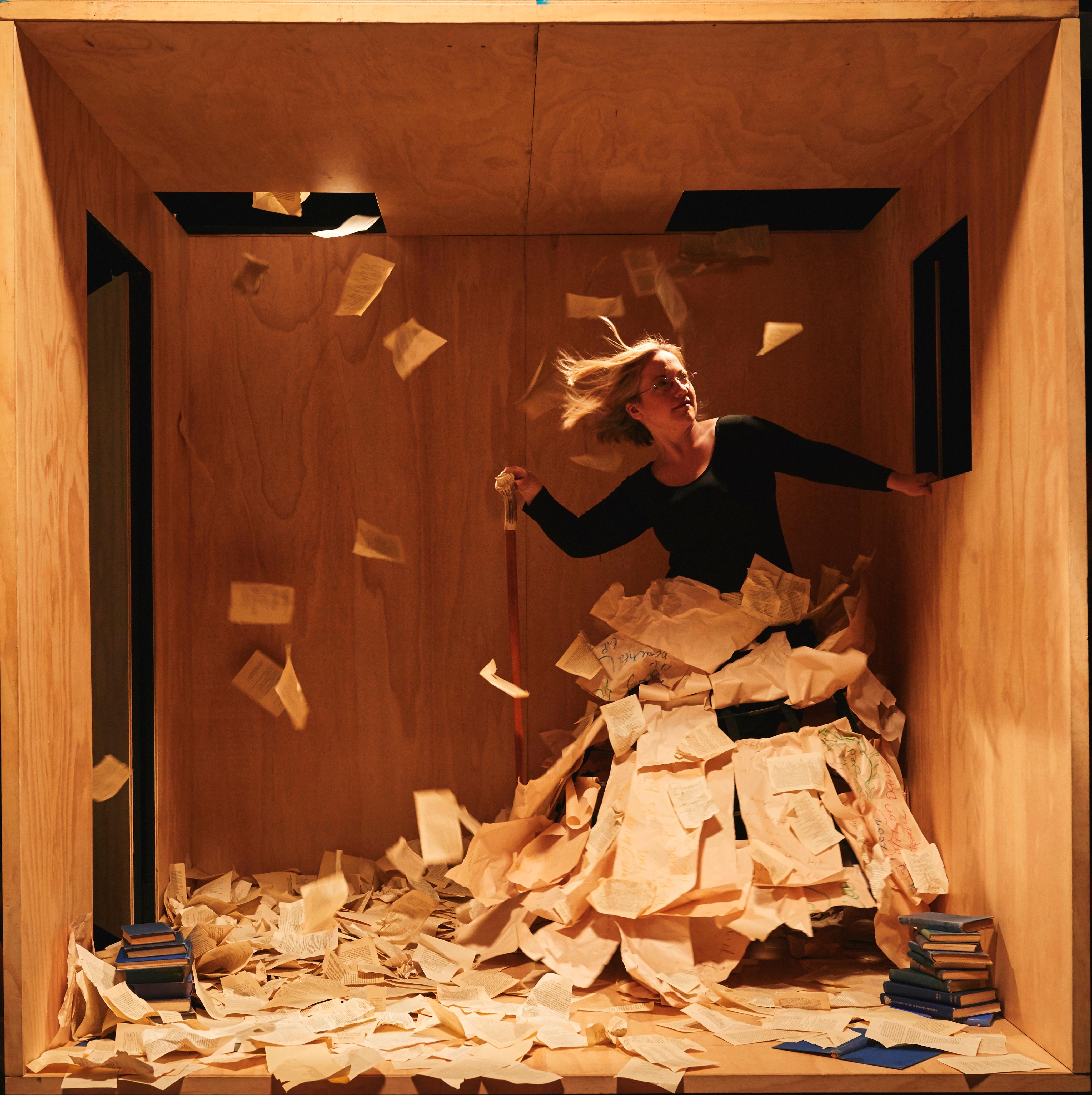 A woman in a large playwood performance box moves to one side. She is covered by book pages falling from large cut-out holes in the ceiling. There are pages all around her.