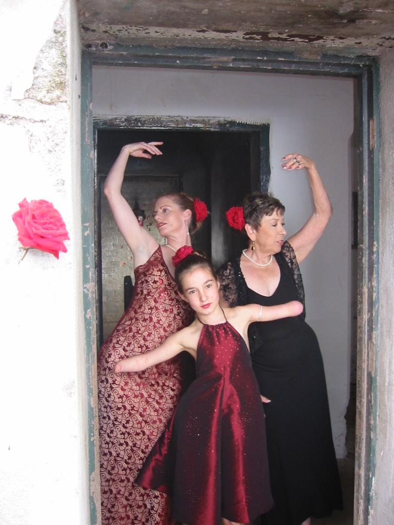 three female-presenting people of different generations, wearing beautiful dresses, with arms extended in a doorway of an old bunker. They each wear a red flower in their hair, looking askance.