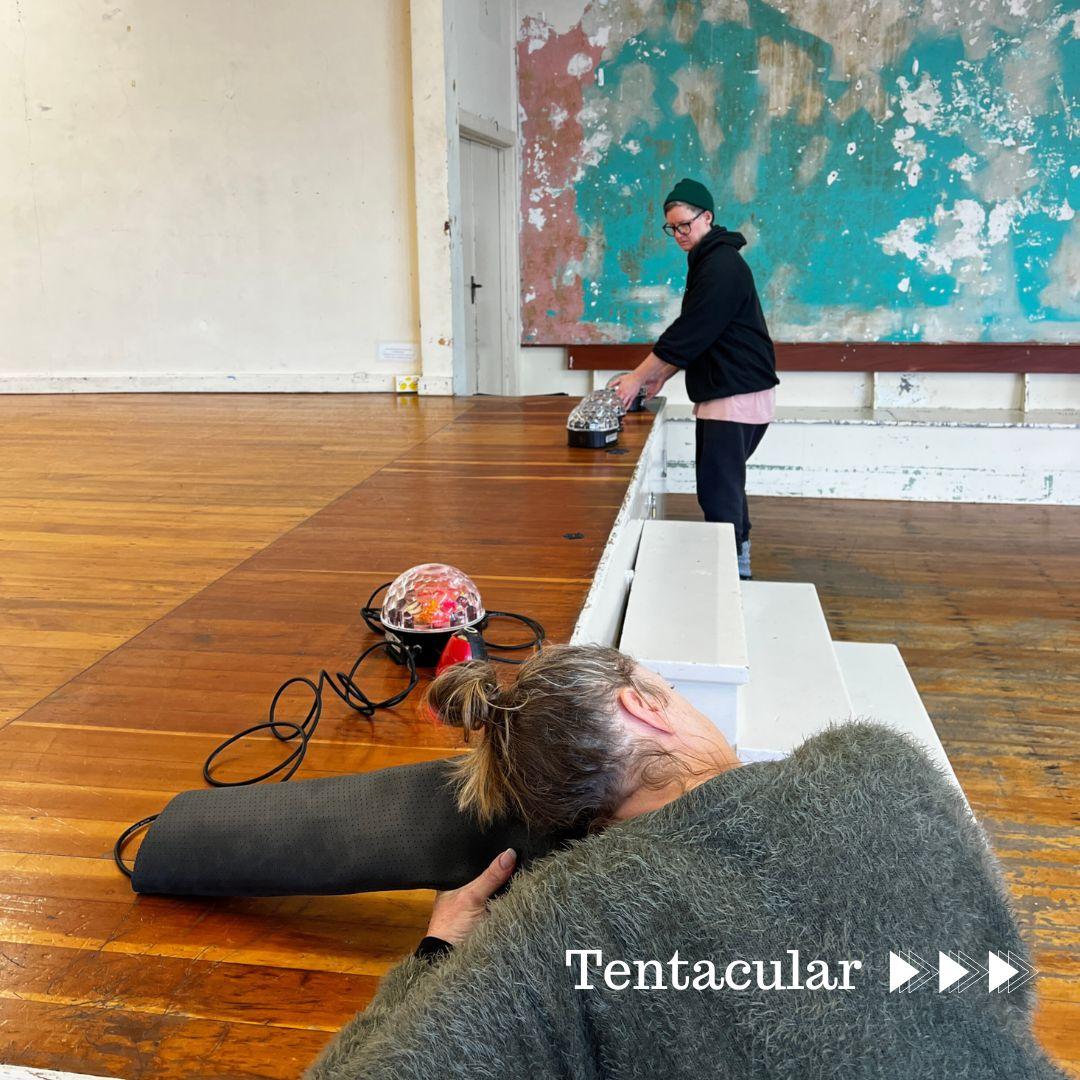 A wooden stage in a worn old hall with peeling greeen and orange paint on the background, two people using the elevated stage to play with and listen to reflective orbs as transducers. One person on the far side of the stage is making sound into one transducer, while the other person, wearing a fuzzy wool sweater and hair up in a bun, is sitting close to the camera, is leaning over, listening to the transducer through a black tube.