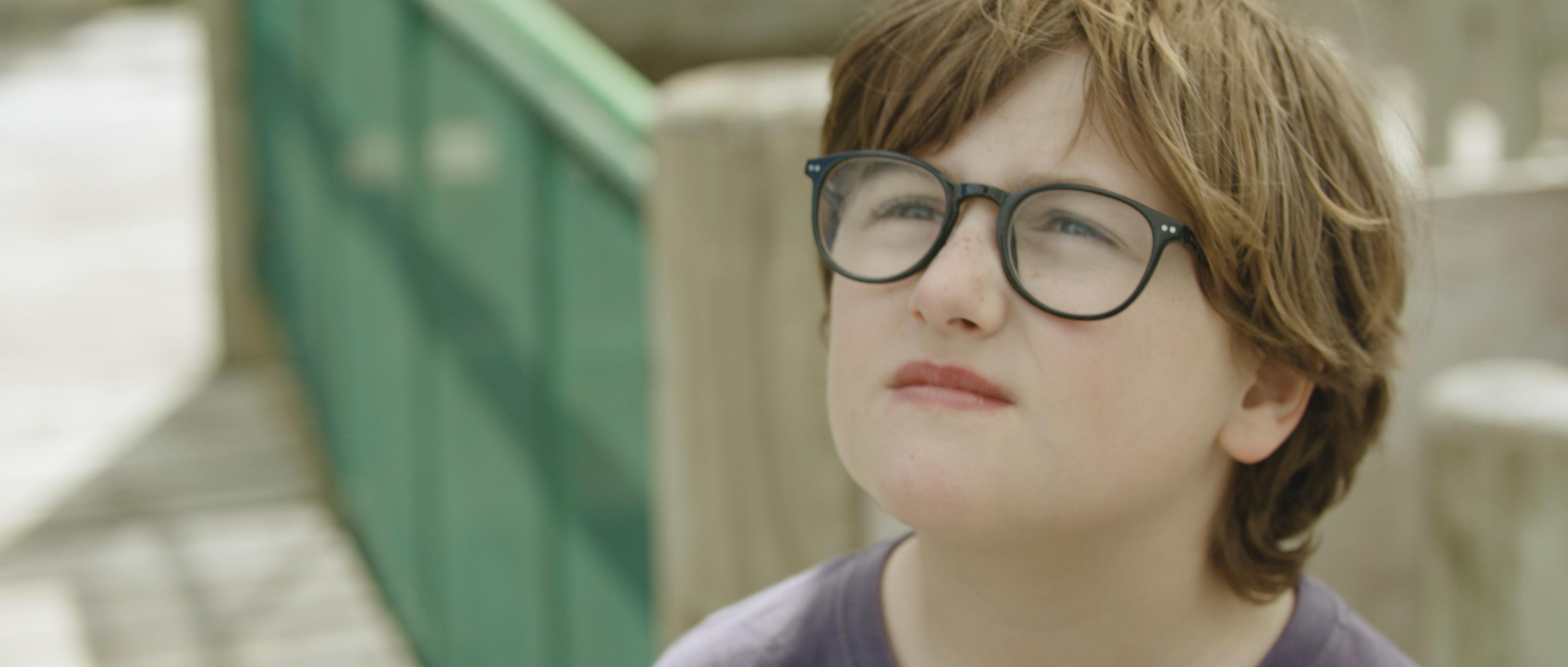 Still from 'Born Different' - a child with light brown short hair, round dark-rimmed glasses and rosy lips looking up towards the sky, on a background of a wooden play structure.