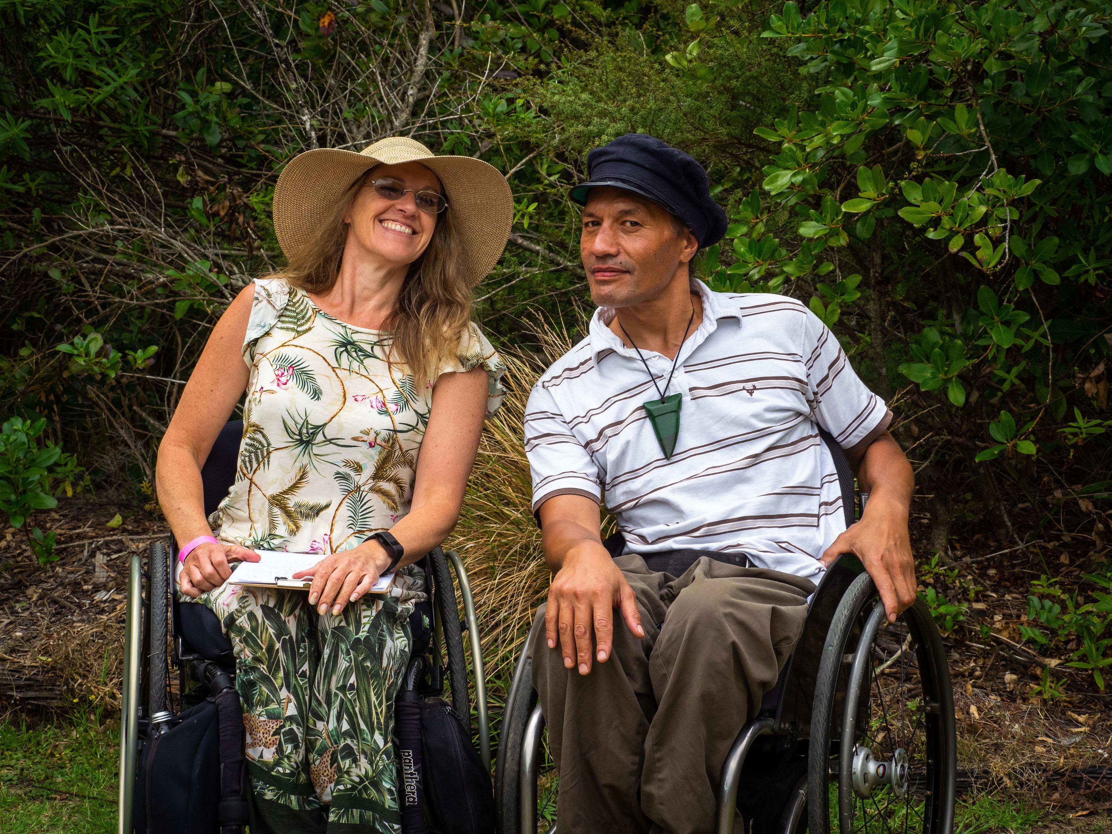 Two people in a natural setting, one light-skinned female, one Māori male, leaning in towards eachother and smiling at the camera.