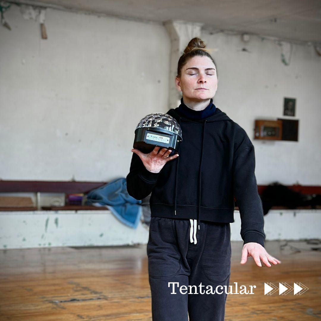 A pale skinned dancer with her hair in a bun, on her head. She is manoeuvering through a an old, aged dance studio with her eyes closed, her left arm out in front of her, her right hand as a platform with an orb as transducer helping her find her way to her destination.