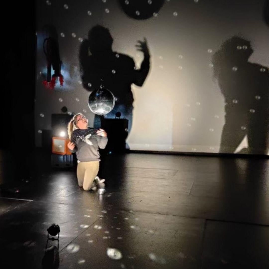 In a dimly-lit room, a woman kneels on the black floor, holding her arms bent upwaards from her waist, and looking at a hanging mirror ball, close to her head. The lighting is directed at the ball, reflecting onto her, and making round hollow dots on the ground and on the wall behind her. The back wall is lit from a different angle, creating two shadows of the dancer, looming large on the wall.