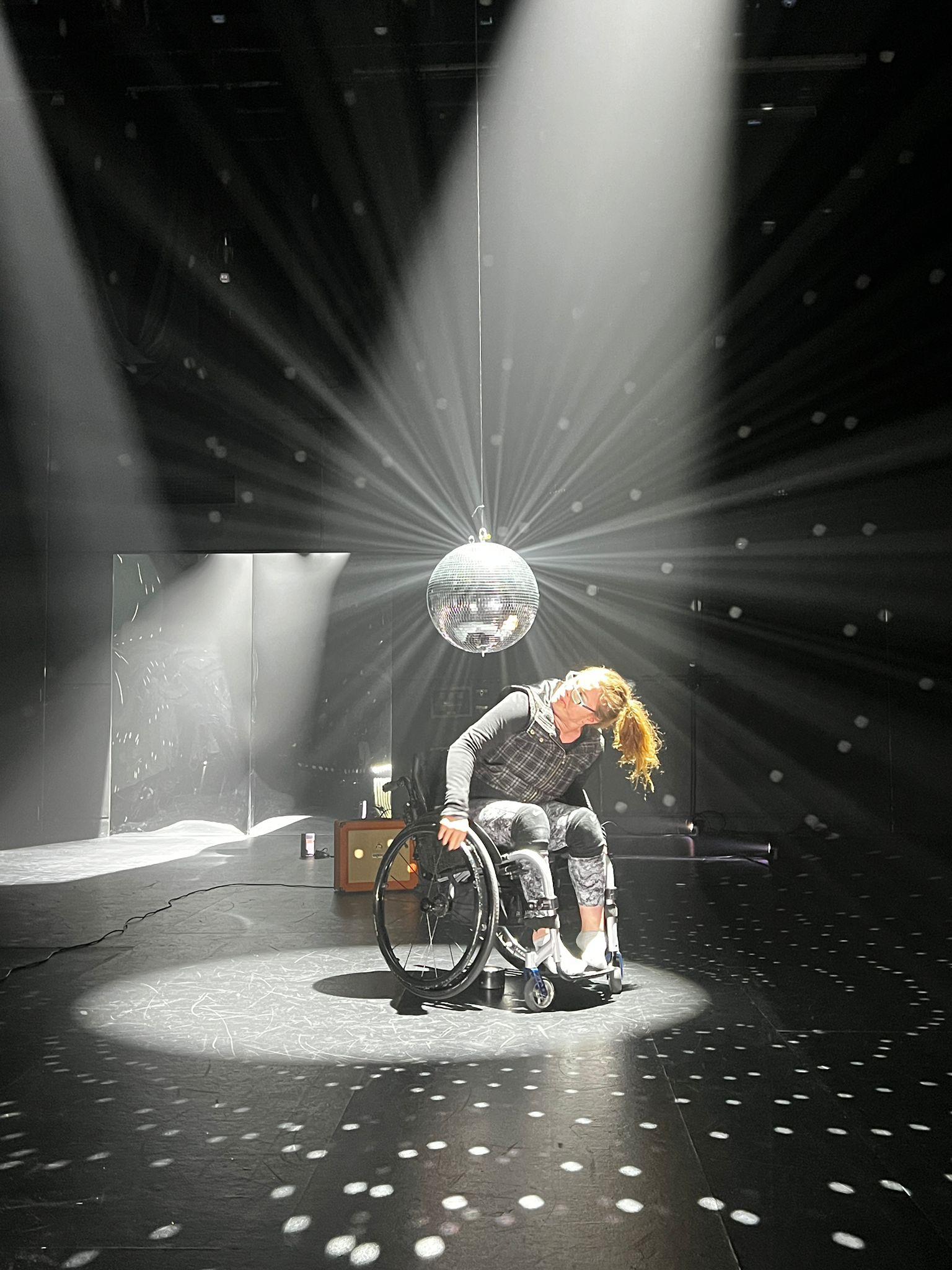 Alisha McLennan Marler, a light-skinned female in a dark top and black and white mottled leggings, sits in a wheelchair in a darkened dance studio, scuffmarks on its black floors, walls black, lights down. She dances around a suspended discoball, didging her head this way and that, as she circles around the base of the ball, watching her reflection. Hundreds of rays of light cut through the hazy room. creating white dots on the walls and floors, in all directions.
