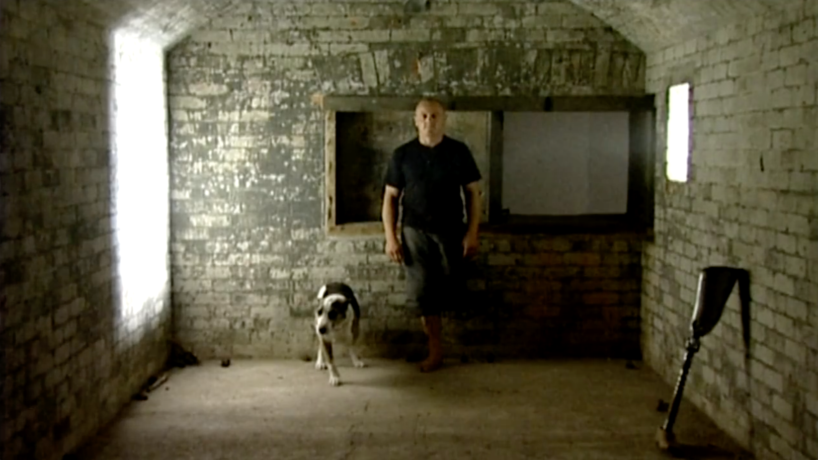 A Man in the back of a world war 2 bunker, next to a three legged dog. The room is brick and dimly lit. They are facing the camera.