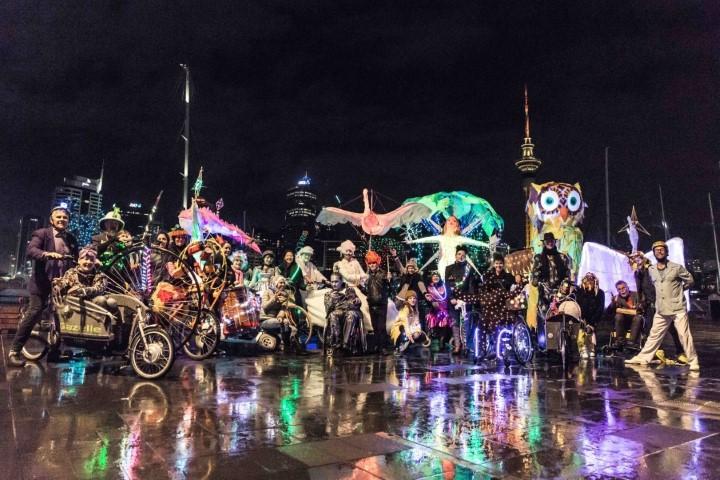 Many costumed people with various kinds of lights on wheels, with wheelchairs, and more, standing on a surface wet with rain
