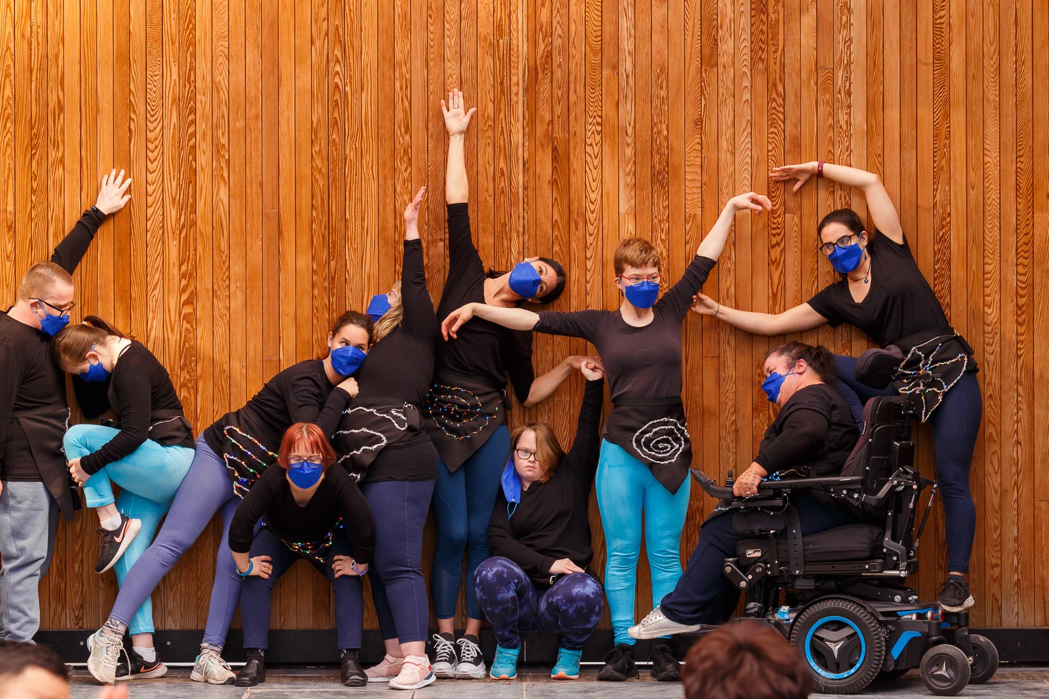 10 dancers, wearing black tops and royal blue face masks, stand close together, making shapes against vertical brown planks of wood.