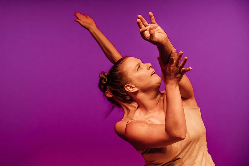 Two dancers in a line, wearing skin-toned clothing, looking to the upper left, with their left hands in the air.