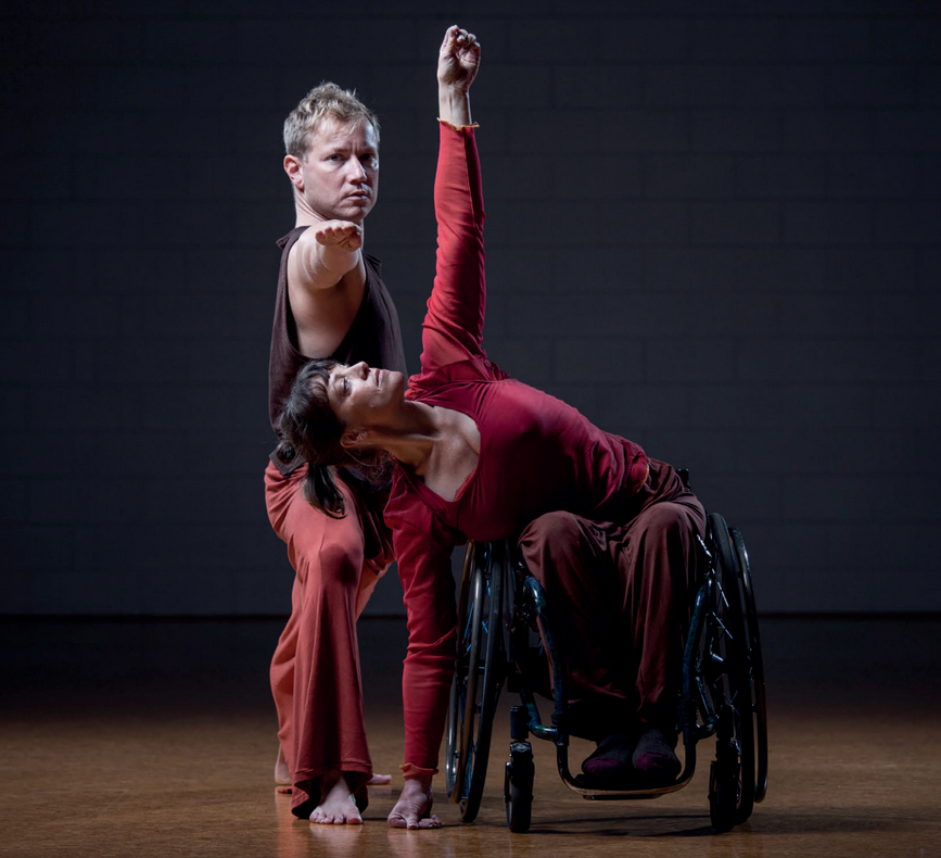 Two dancers in a dark room. One is on a knee, arm straight in front, looking straight at the camera. The other is in a wheelchair with arms stretched from floor to ceiling, looking at the ceiling.