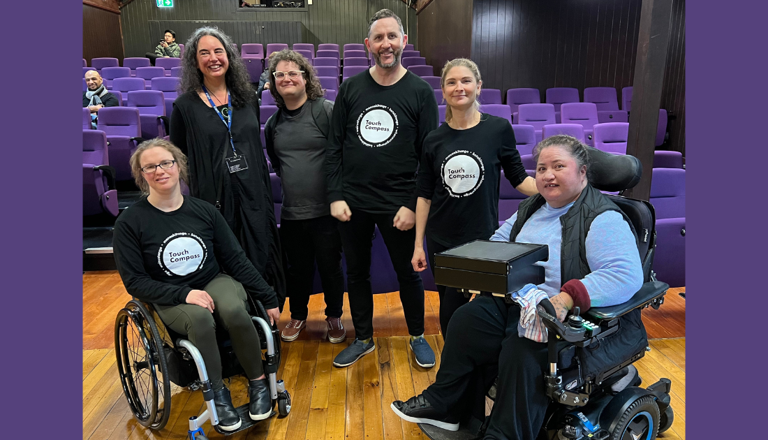 Six people in a semi-circle, two in wheelchairs flanking four people standing, in the centre, in a brightly-lit performance theatre. Three people are wearing black shirts with a white Touch Compass logo. They are posing with wide grins for the camera.