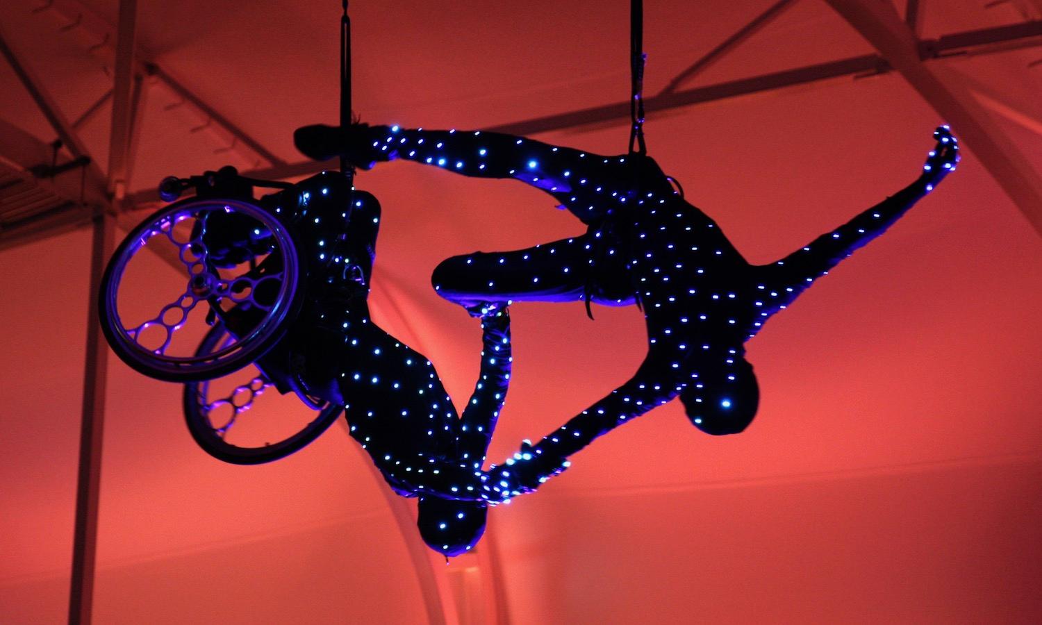 Two Aerial dancers in black clothing, covered with LED lights, in the air against a red background, in a tent. One is in a wheelchair, the other is not.