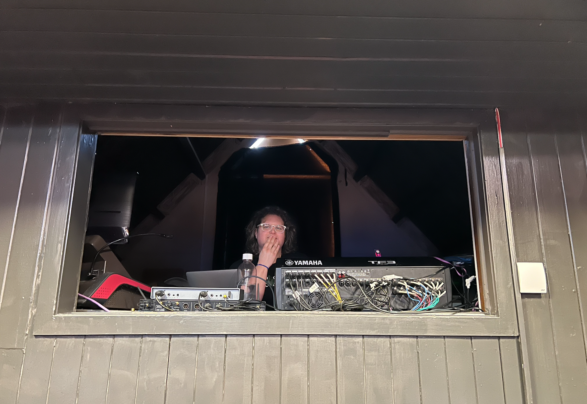 Creative Producer Izzy Robinson (they/them) sits in the control room at the back of an old theatre. They are weaing white round glasses, their right hand on their chin, sitting behind adio visual controls, with lots of cables and switches.