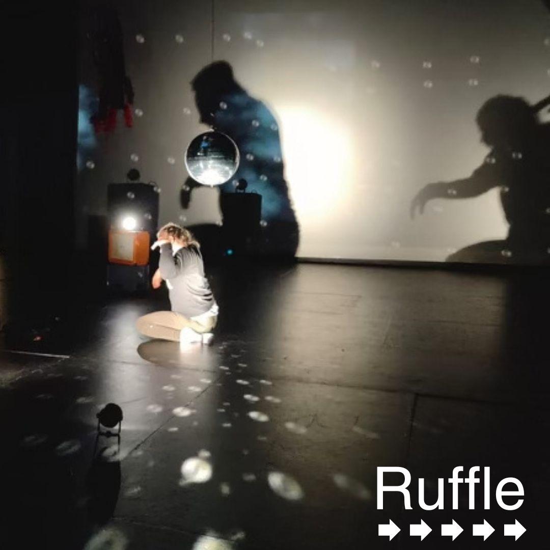 In a dimly-lit room, a woman kneels on the black floor, holding her arms bent upwards from her side. She is perpendicular to the rear wall, underneath a hanging mirror ball, close to her head. The lighting is directed at the ball, reflecting onto her, and making round hollow dots on the ground and on the wall behind her. The back wall is lit from a different angle, creating two shadows of the dancer, looming large on the wall.