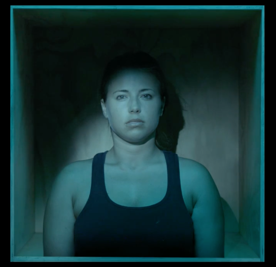 A woman with hair tied back and a dark singlet stands against a wooden wall. She is dimly illuminated by blue light. She looks enclosed, almost uncomfortable.