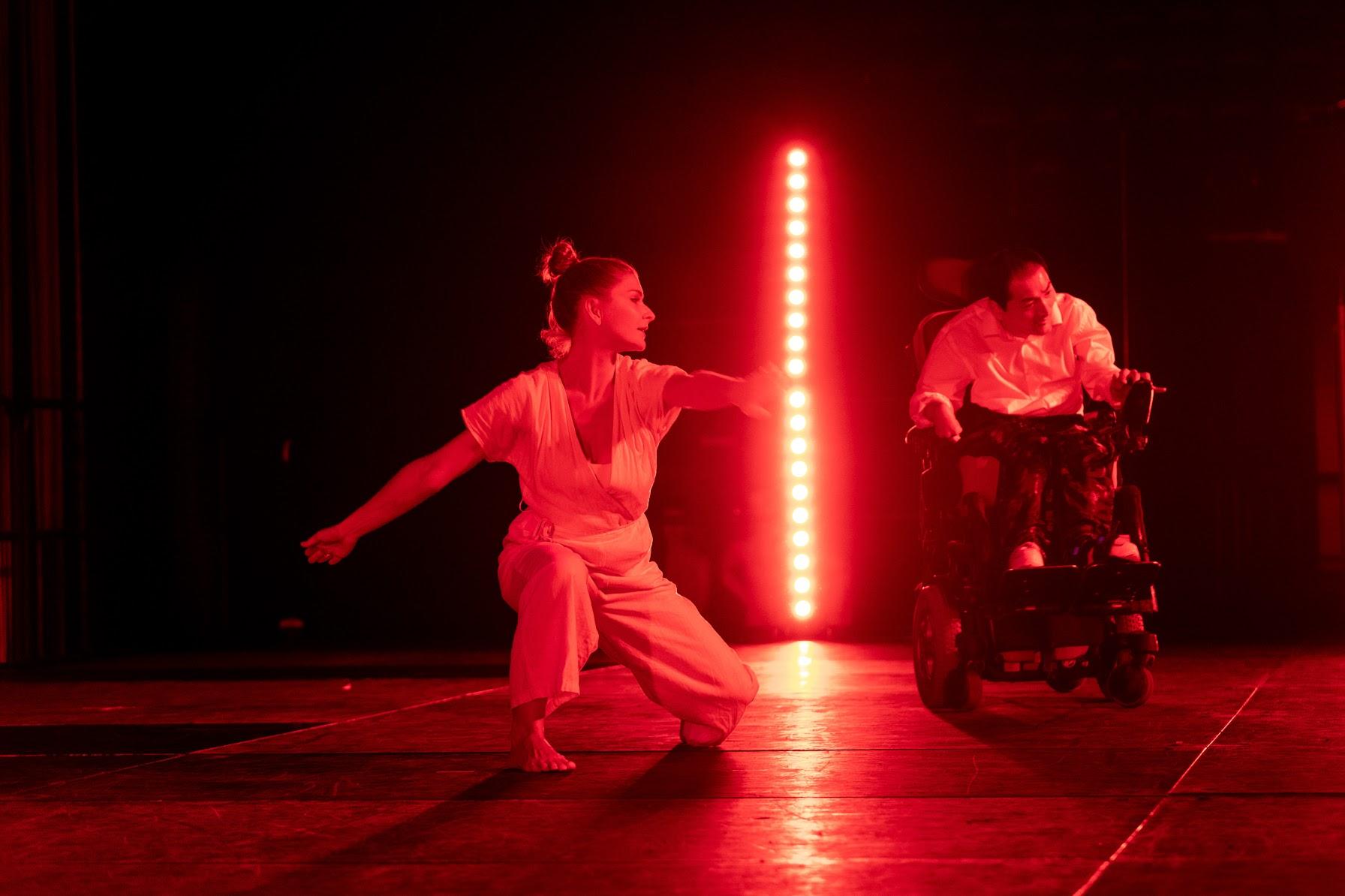 Two people dancing on stage: a performer in a powerchair, and a dancer in a white costume on her knees; a string of bright verticle LED lights separates them.