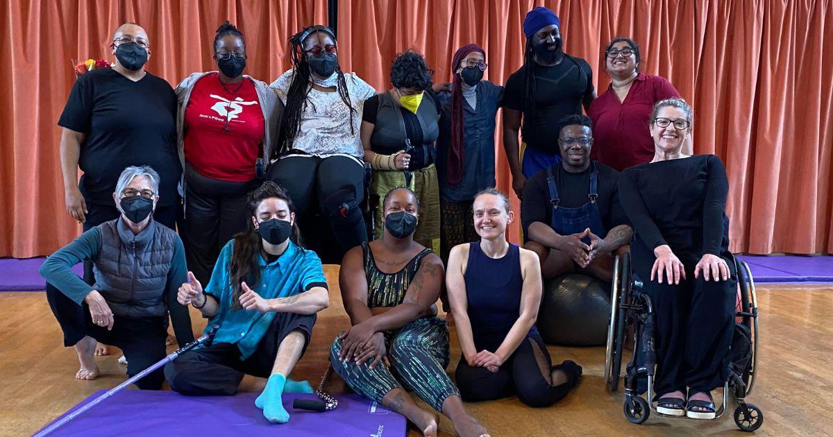 A group of 13 disabled dancers with different ethnicities standing and sitting in two rows. Many of them wear facemasks. They all look at the camera smiling at the thrill of being part of the workshop.
