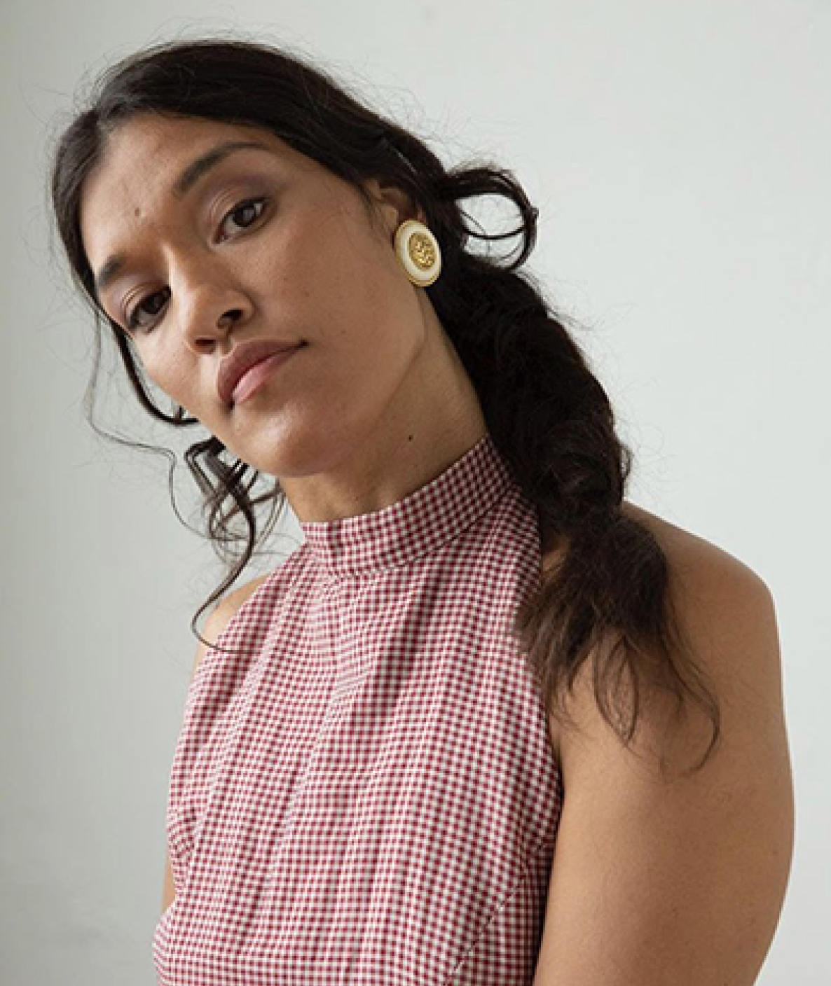 A tan-skinned woman with long dark hair in a ponytail, head cocked to the side, wearing a checked sleeveless shirt, looking at the camera.