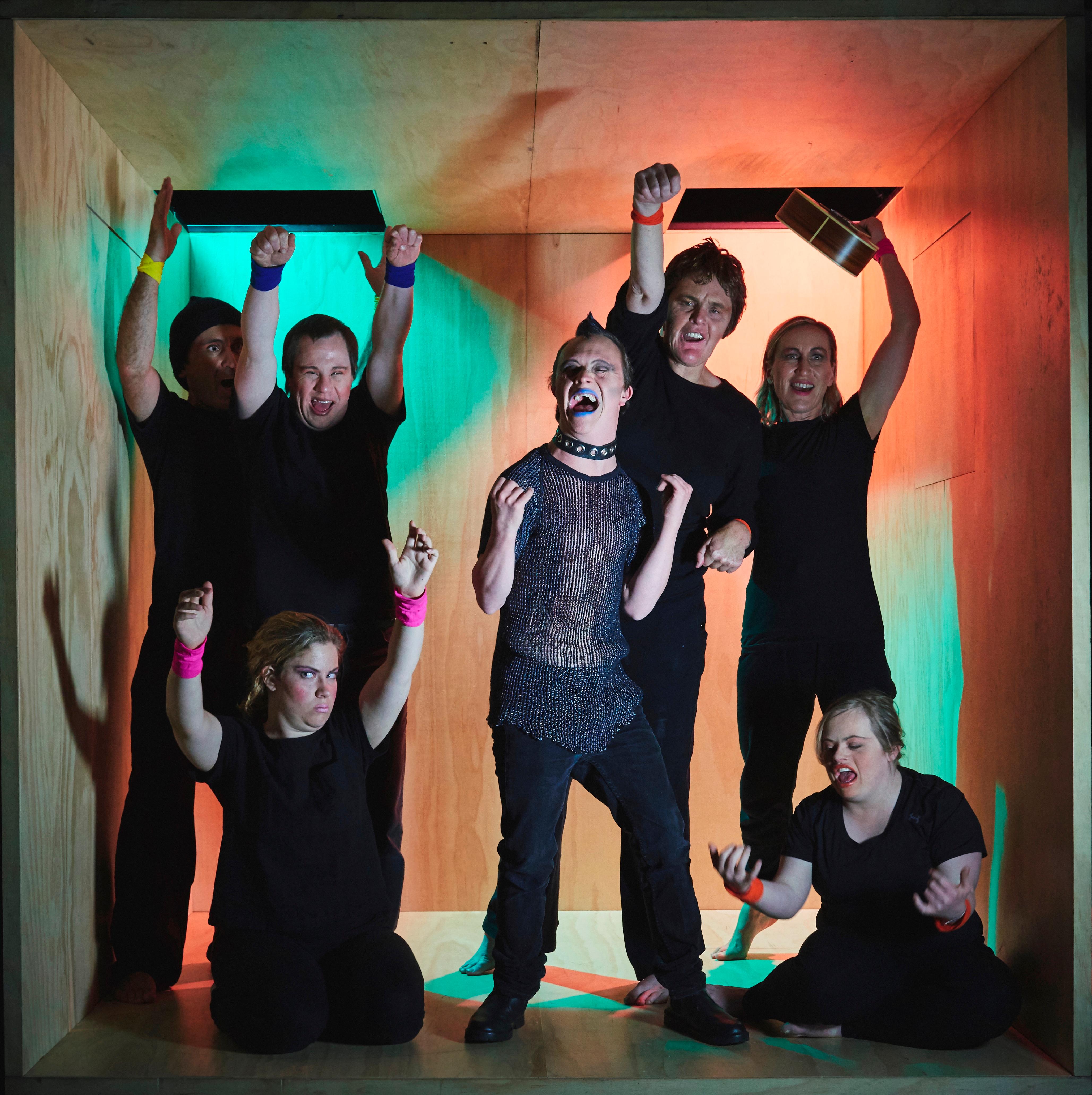 Seven people wearing dark clothing with neon armbands around their wrists yelling in unison. They stand in a large wooden performance box, around a mane with a sharp dog collar and spiky hair, looking punk rock!