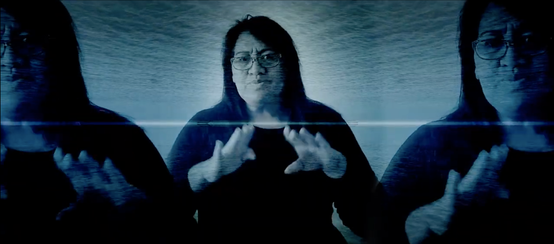 A still from Alter State Digital Launch. A Māori female sign language interpreter with long dark hair and glasses, wearing a dark shirt, is set against a background of a top-to-bottom mirror image of ocean ripples. She is using her hands to interpret.