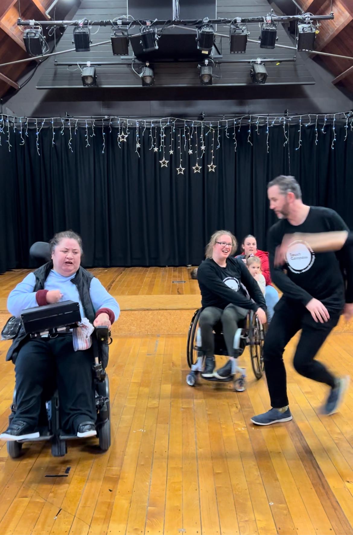 Three Touch Compass dancers in a theatre, dancing. One dancer is in a powerchair, another in a wheelchair, and the third standing and dancing. They are improvising to the dance, led by Touch Compass dancers.