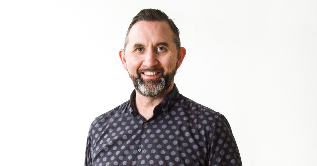 A light-skinned Māori man with energetic eyes framed by heavy eyebrows, wearing a salt and pepper beard, with short cropped hair and a blue/black polkadot shirt. He is smiling.