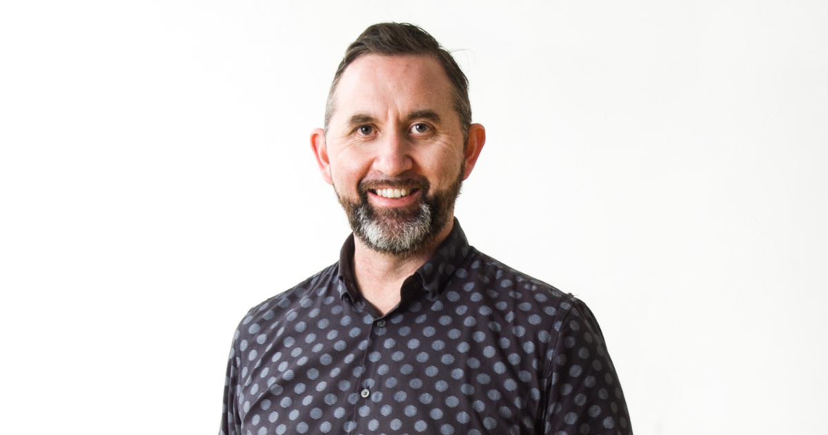 A light-skinned Māori man with energetic eyes framed by heavy eyebrows, wearing a salt and pepper beard, with short cropped hair and a  blue/black polkadot shirt. He is smiling.