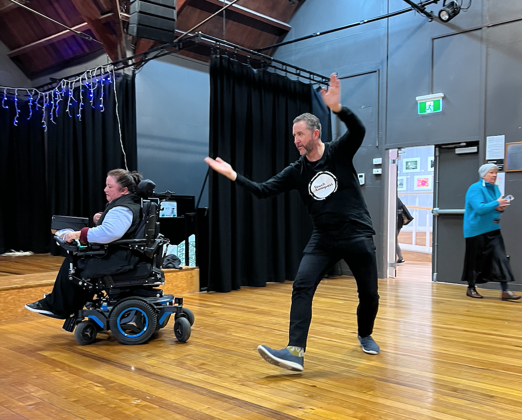 Two dancers in a room with a reflective wood floor; one dancer in a wheelchair, the other dancing on his feet, hands in the air, moving to one side. They both wear black trousers and are intent on their movement.