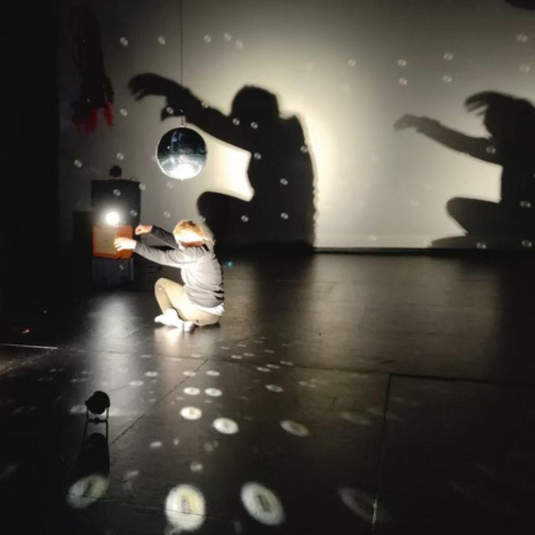 In a dimly-lit room, a woman kneels on the black floor, holding her arms bent upwards from her side. She is perpendicular to the rear wall, underneath a hanging mirror ball, close to her head. The lighting is directed at the ball, reflecting onto her, and making round hollow dots on the ground and on the wall behind her. The back wall is lit from a different angle, creating two shadows of the dancer, looming large on the wall.