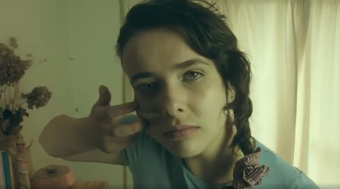 A girl with long dark hair in a braid to the left side of her hear smears grey face paint in two marks across her face. She looks at the camera, with a determined look. She is in a dark room with dried flowers.