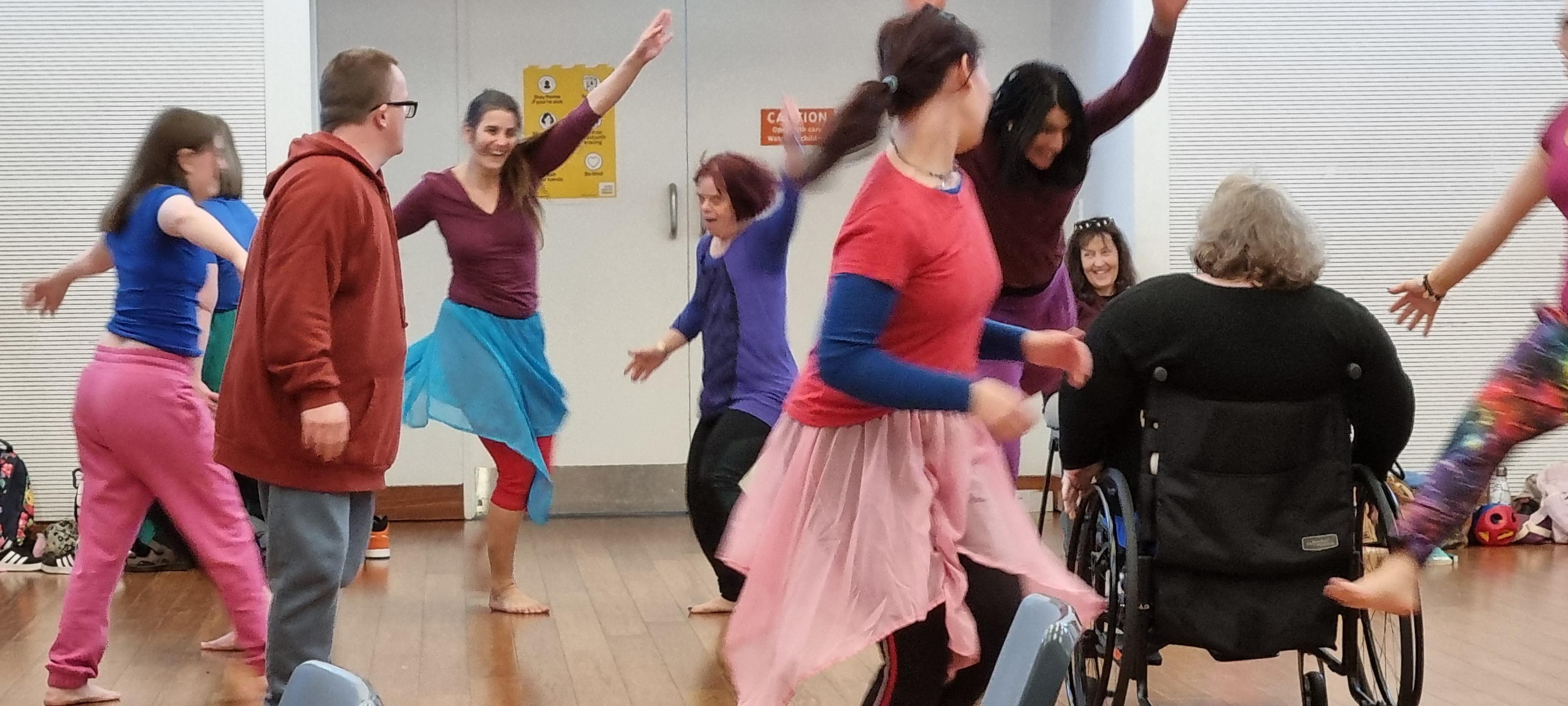 The Touch Compass Contemporary Class. Many people wearing block coloured clothing dancing at the same time, most of them standing, one person in a wheelchair. Some have their hands in the air. They are enjoying themselves.