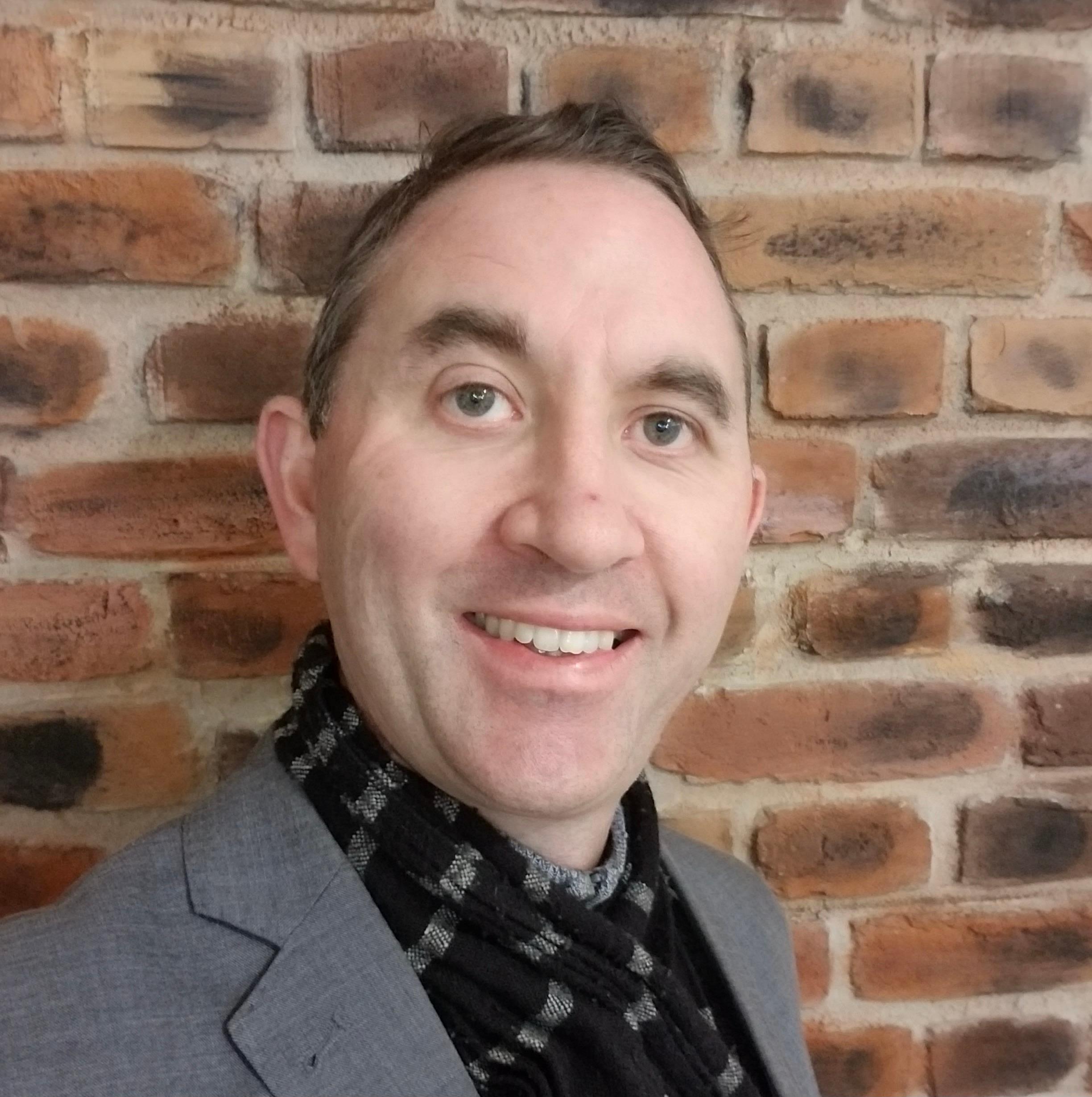 A light-skinned Māori man with short dark hair and bushy eyebrows against a brick wall, wearing a jacket and scarf, smiling at the camera.