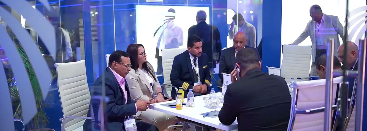 Delegates, one of them a pilot in a captain's uniform, meet around a white table inside a glass-walled stand at EIAS 2026