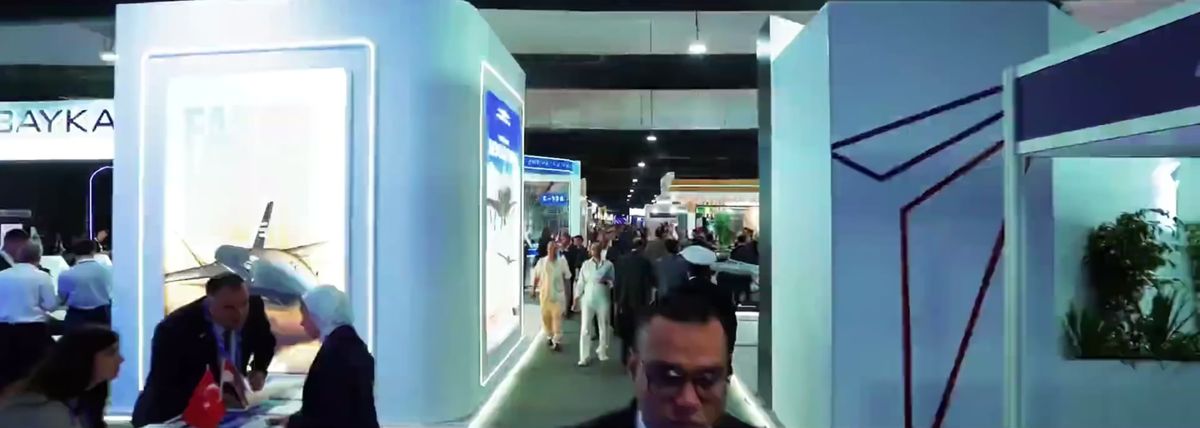 Visitors fill a long aisle between white, LED-lit stands at EIAS 2026, a man in glasses walking toward the camera