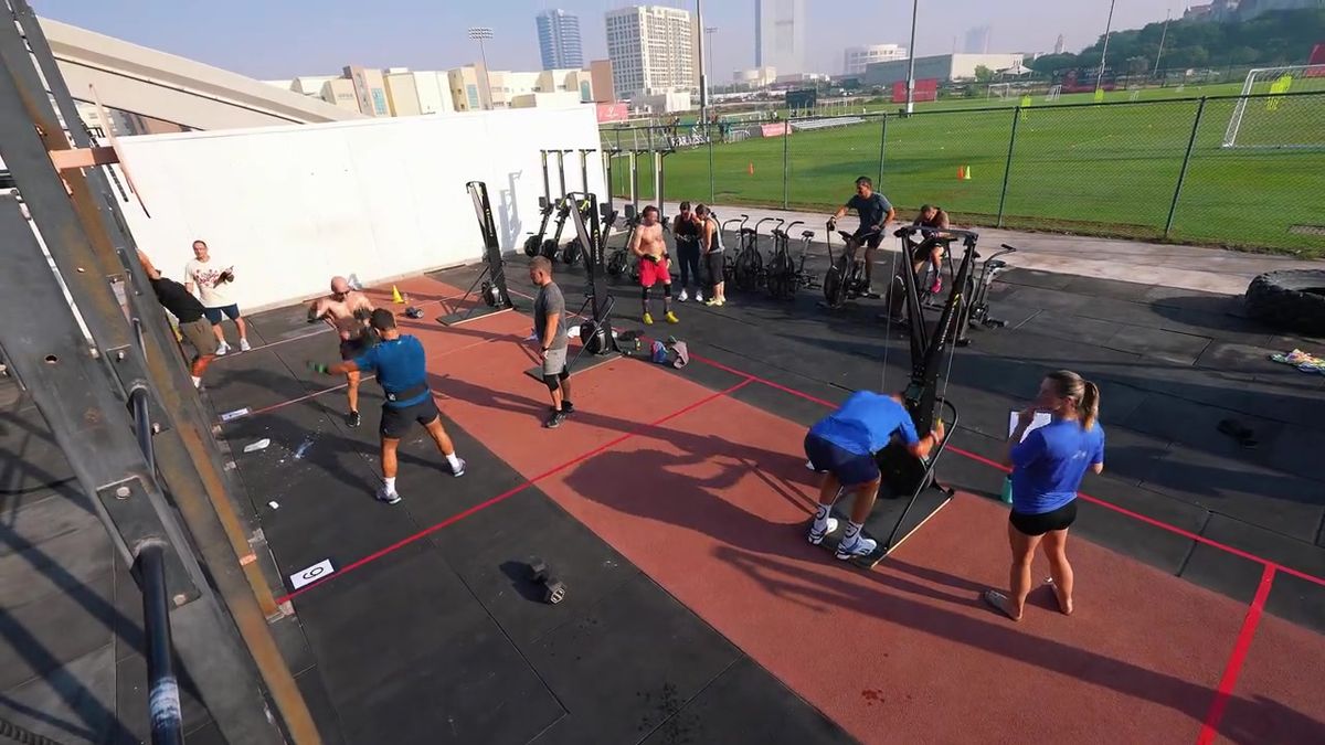 High-angle view of an outdoor CrossFit competition in Dubai: athletes on ski ergs and air bikes, a judge with a clipboard, a football pitch and towers behind