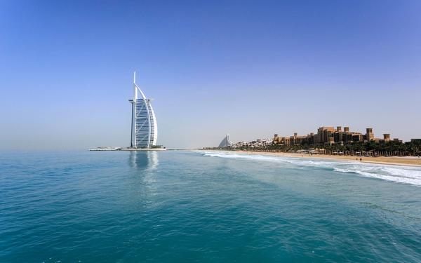 Burj Al Arab rising from the sea beside a long Dubai beach, under a clear blue sky