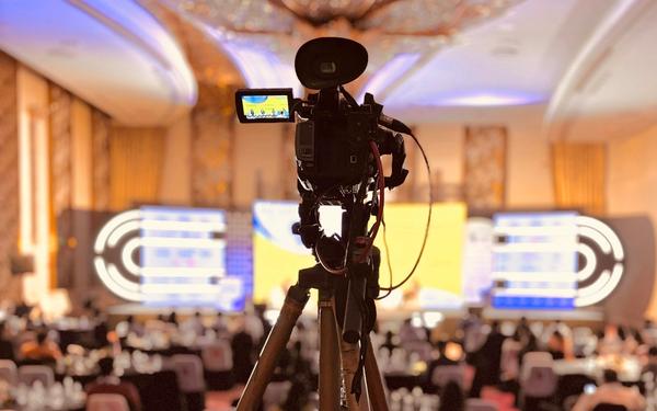 Video camera on a tripod at the back of a conference ballroom, with the stage screens in the background