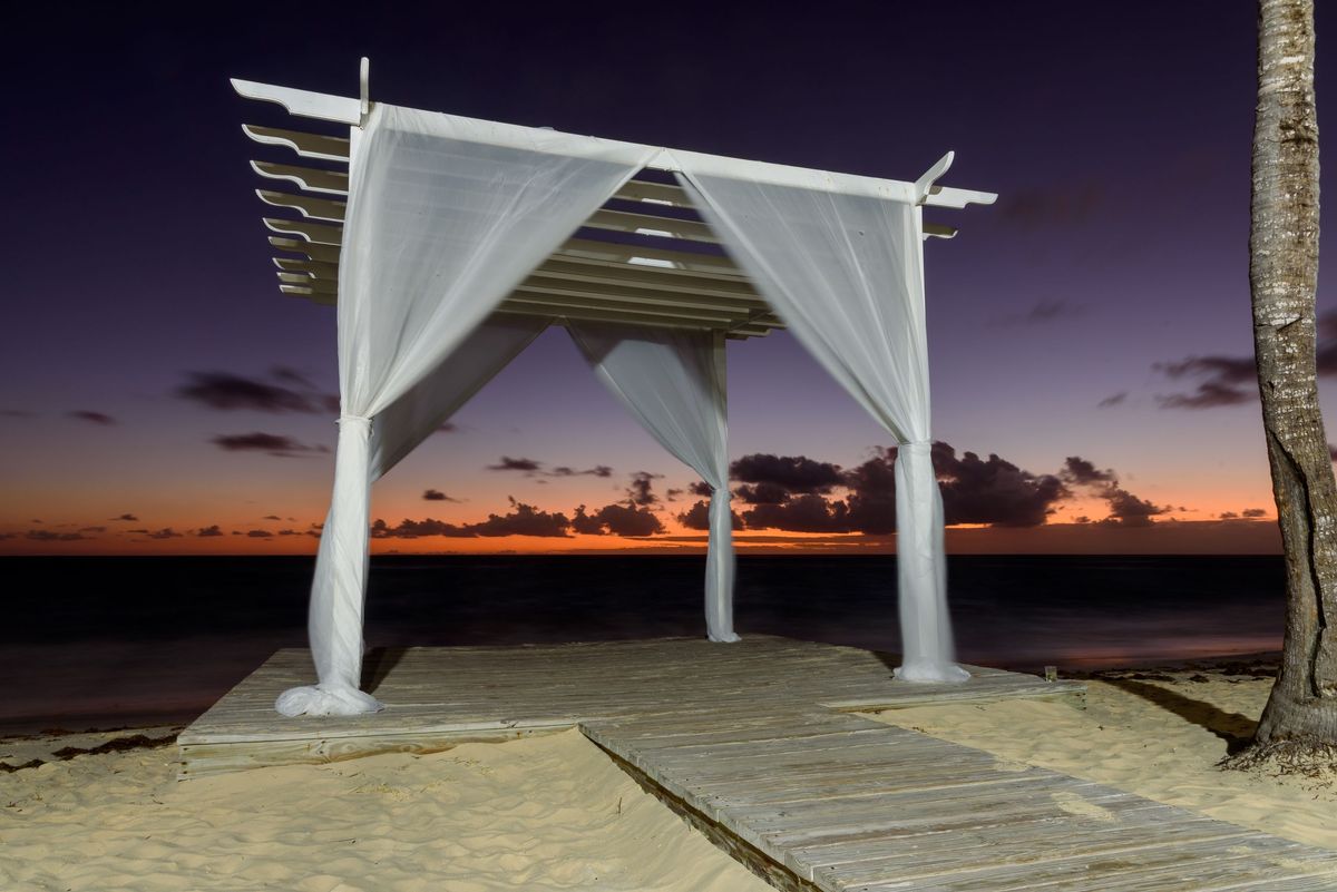 Un pavillon de mariage en bois blanc drapé de voilages, sur un ponton de plage juste après le coucher du soleil, devant la mer et un horizon orangé