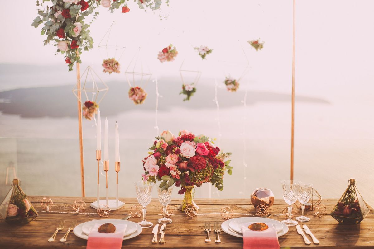 Une table de mariage dressée pour deux, avec des roses, des verres en cristal, de hautes bougies blanches et des fleurs suspendues, la mer en fond dans une lumière douce