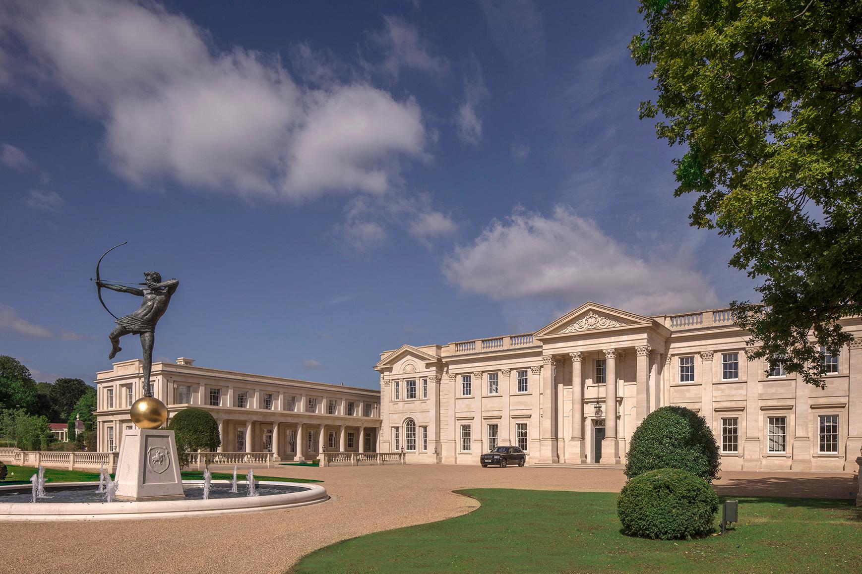 Front exterior of mansion house constructed in natural stone, with sweeping gravel driveway and a circular fountain with an archer sculpture