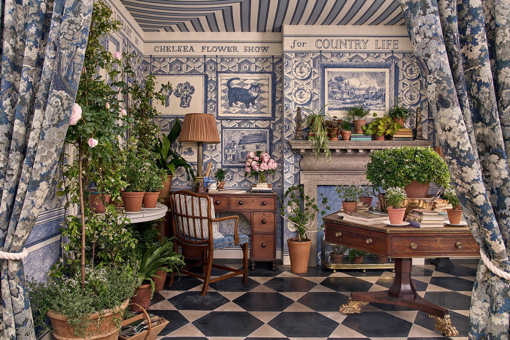 Country Life stand at the Chelsea Flower Show featuring hand-drawn blue and white wallpaper, a classical interior setting with desk, armchair, stone fireplace and potted plants.