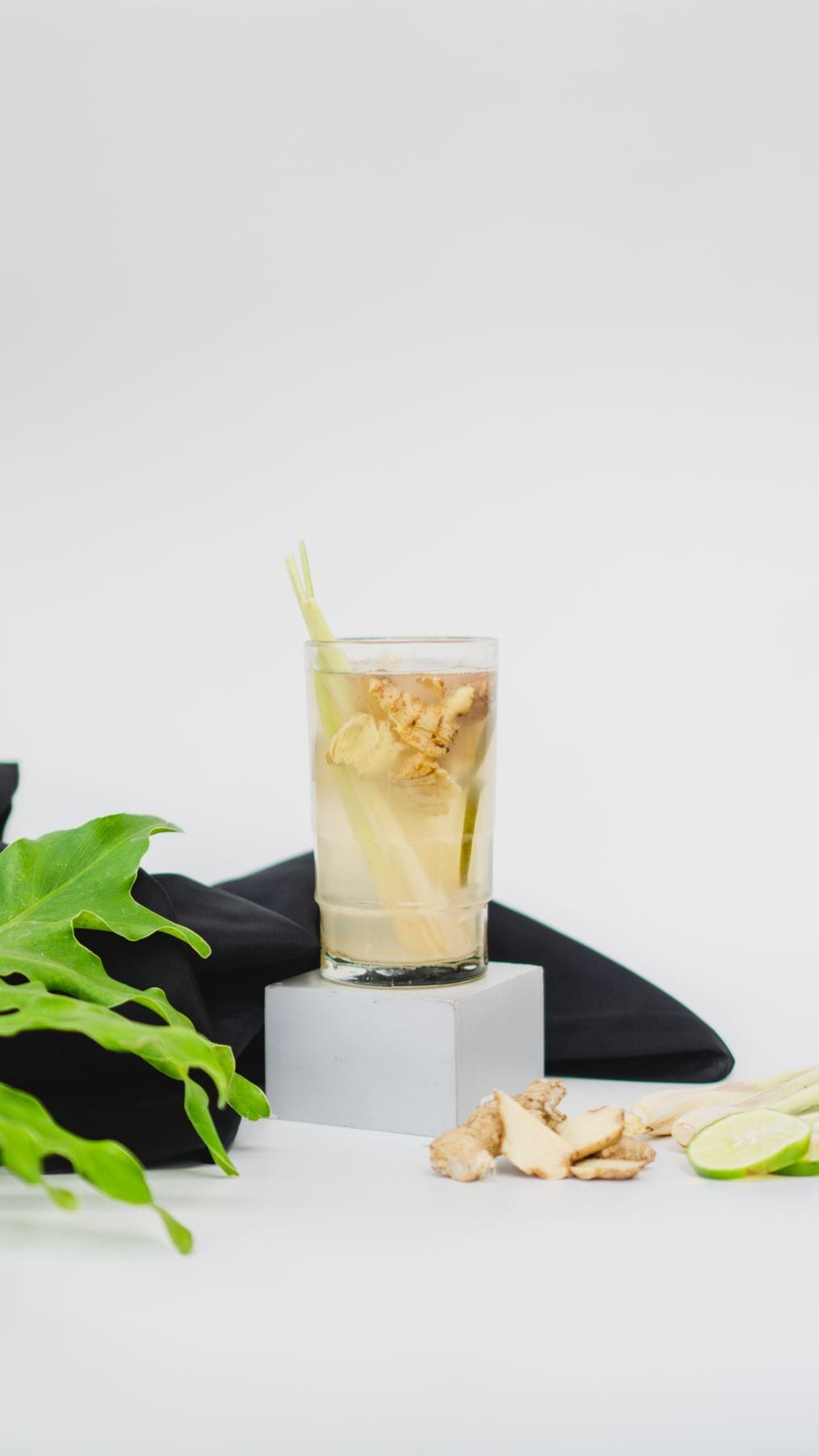 A glass of pale golden ginger brew with sliced ginger and lemongrass inside, fresh ginger root and lime wheels beside it.