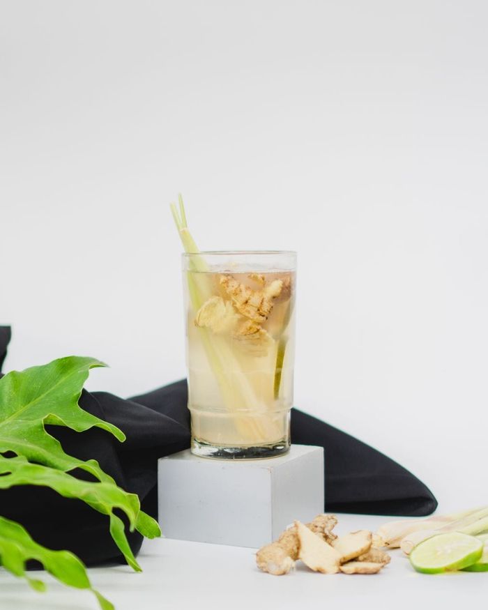 A glass of pale golden ginger brew with sliced ginger and lemongrass inside, fresh ginger root and lime wheels beside it.