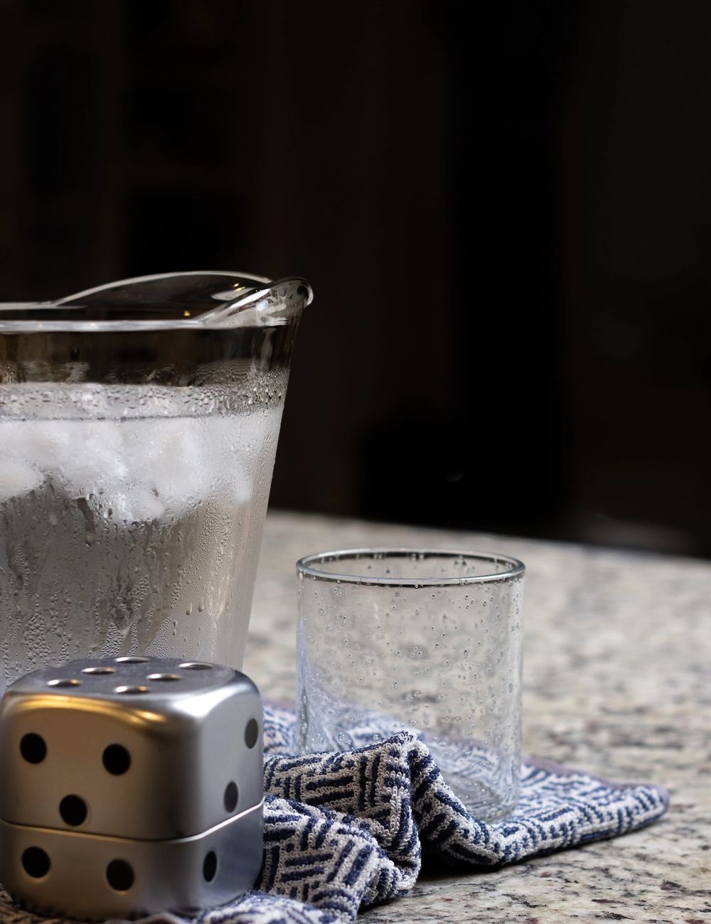 Glass pitcher of iced citrus water beside a tall textured tumbler, a small dice-shaped salt shaker resting on a folded indigo-stripe linen napkin in the foreground.