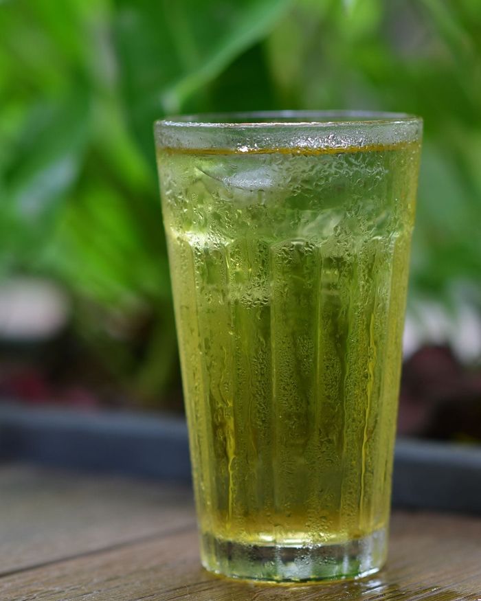 A tall faceted glass of chilled yellow green sencha tea beaded with condensation, lush leaves behind.