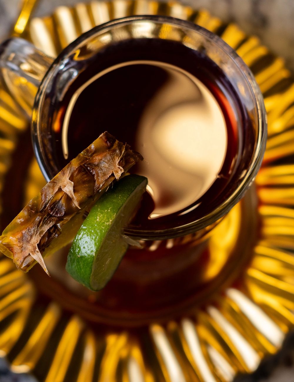 Overhead shot of a faceted glass mug holding deep ruby tea, garnished with a piece of dried ginger root and a lime wedge, on a gold-rayed scalloped saucer.