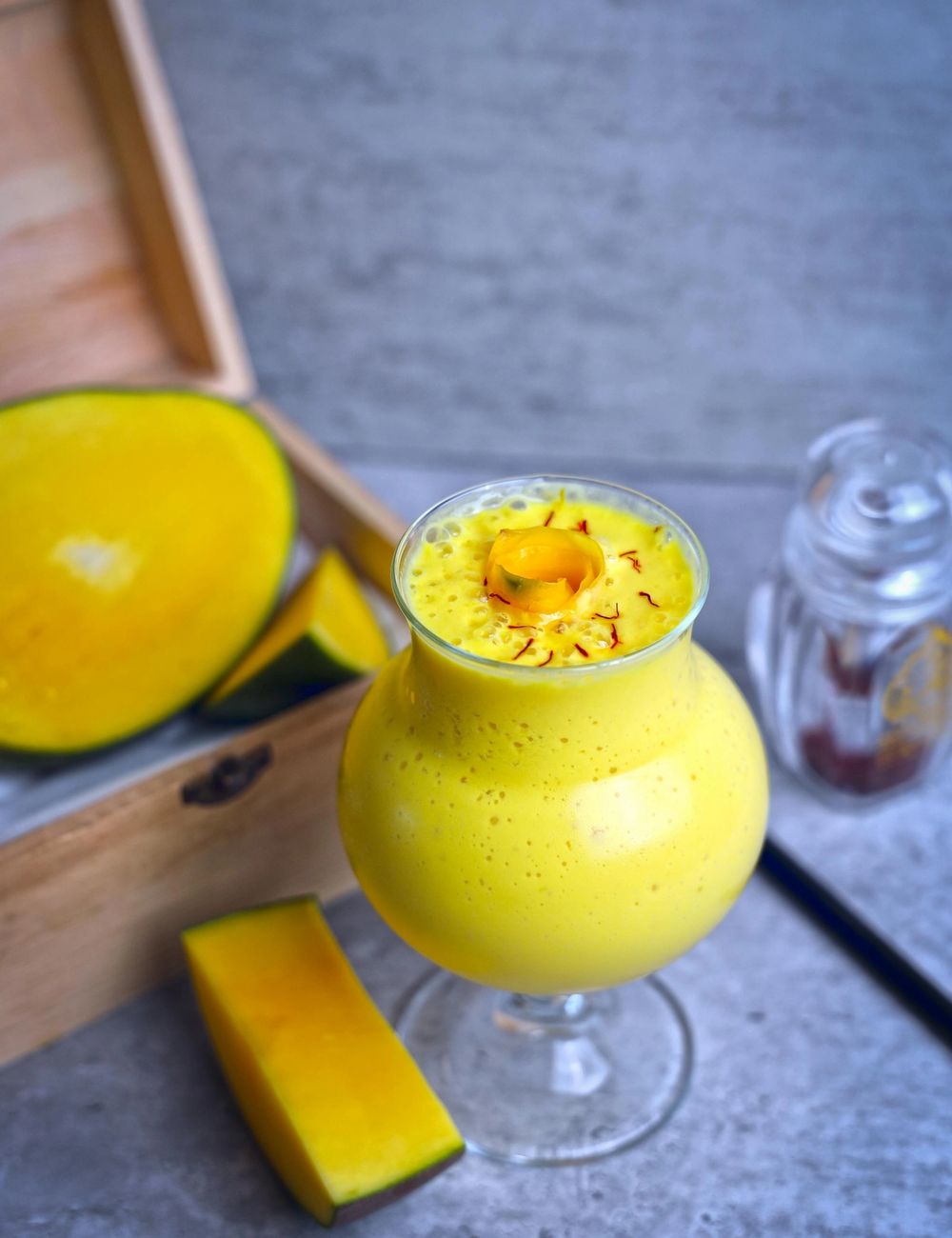 Stemmed glass of bright golden mango lassi, saffron strands trailing across the foam top, mango slices arranged on a small wooden box beside the glass on a soft grey backdrop.