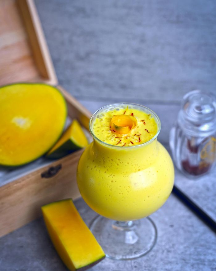 Stemmed glass of bright golden mango lassi, saffron strands trailing across the foam top, mango slices arranged on a small wooden box beside the glass on a soft grey backdrop.