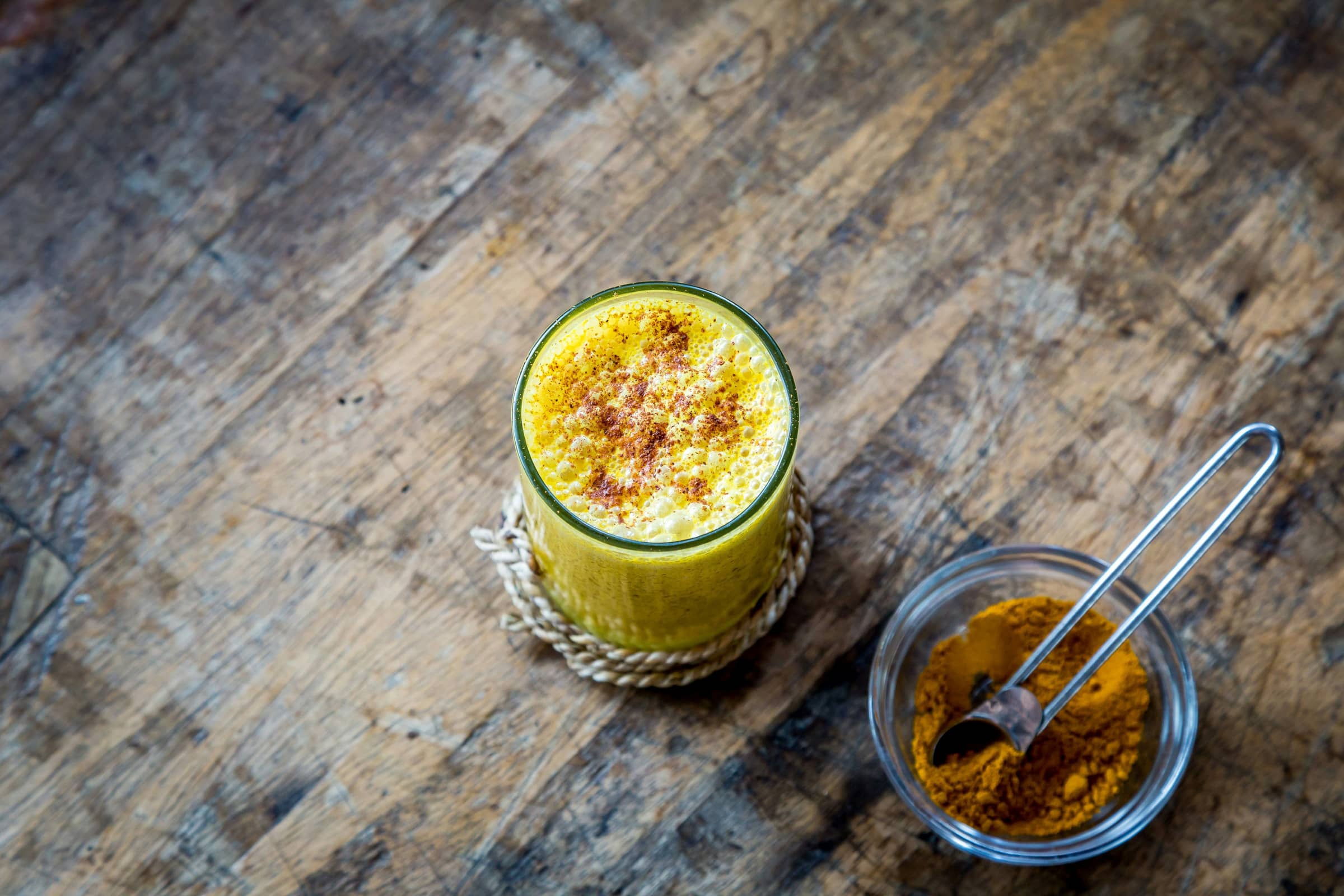 A glass of golden turmeric drink with a frothy spiced top, beside a small bowl of ground turmeric on rustic wood.