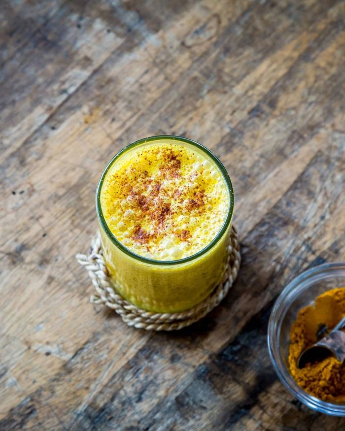 A glass of golden turmeric drink with a frothy spiced top, beside a small bowl of ground turmeric on rustic wood.
